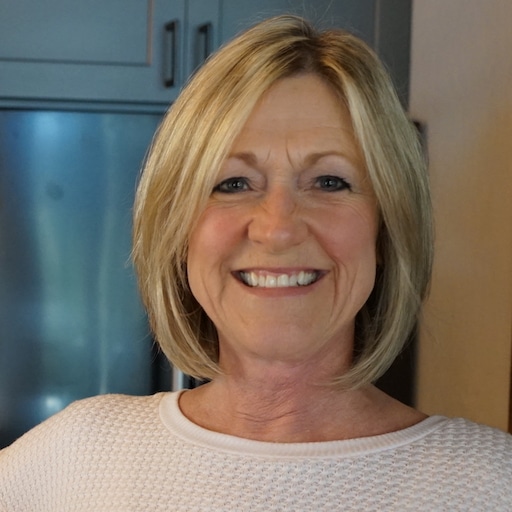 Smiling woman with blonde hair standing indoors with a softly blurred kitchen background.