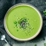 Bowl of creamy green soup garnished with fresh herbs and seeds on a dark cloth with a spoon nearby.