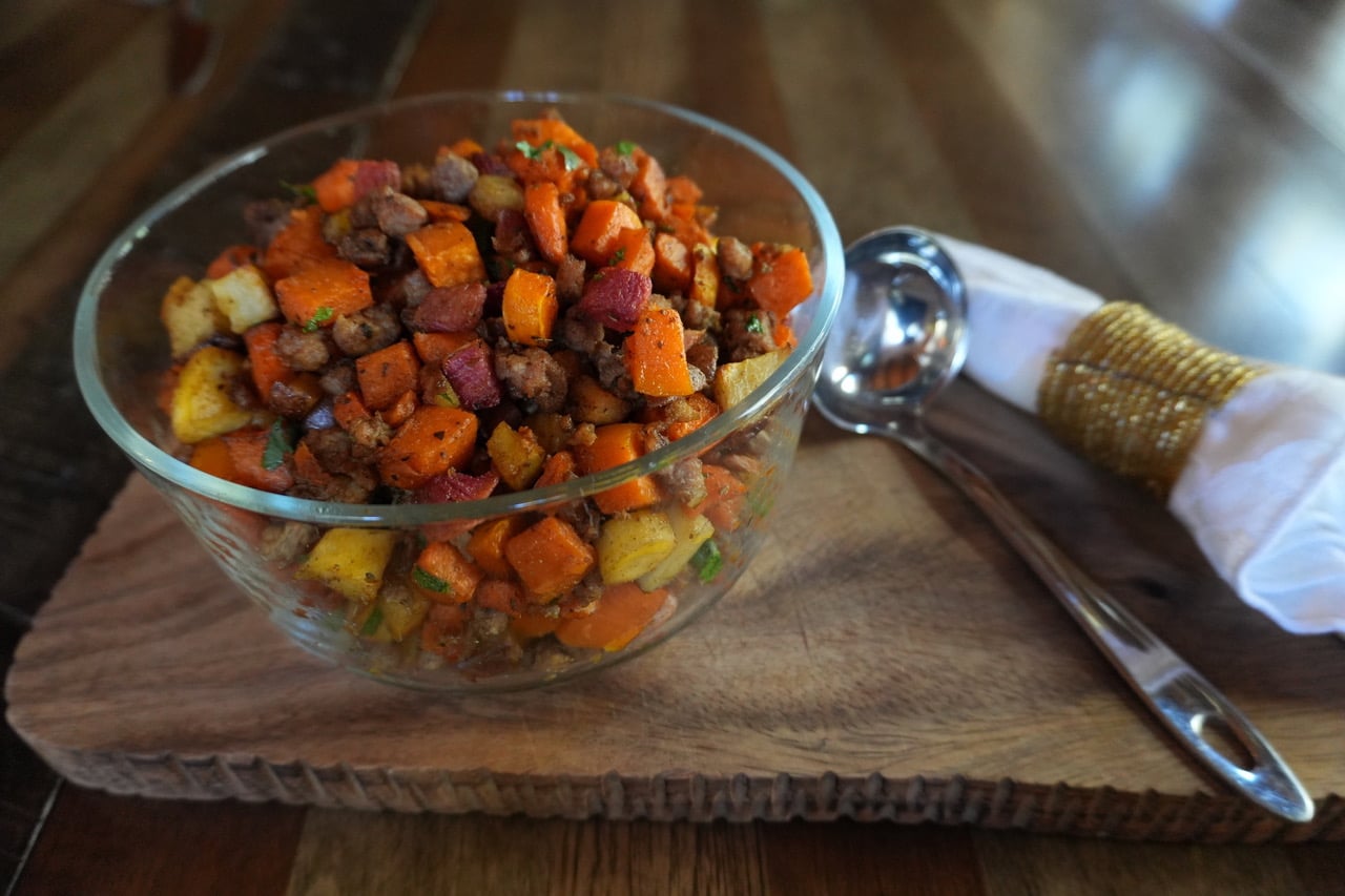 Glass bowl filled with roasted vegetables on a wooden table with a serving spoon nearby.